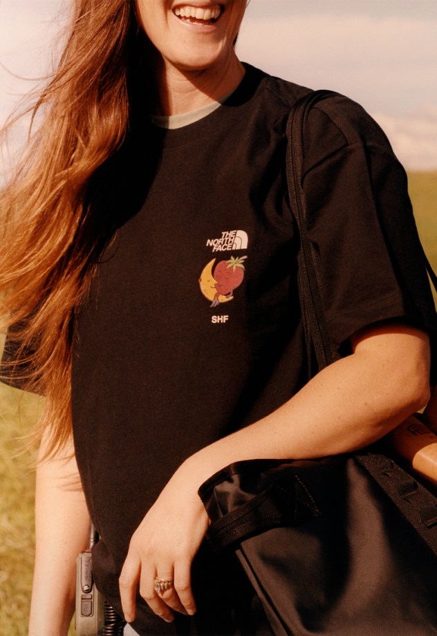 A woman smiles wearing her The North Face x Sky High Farm Goods tee and tote bag. 