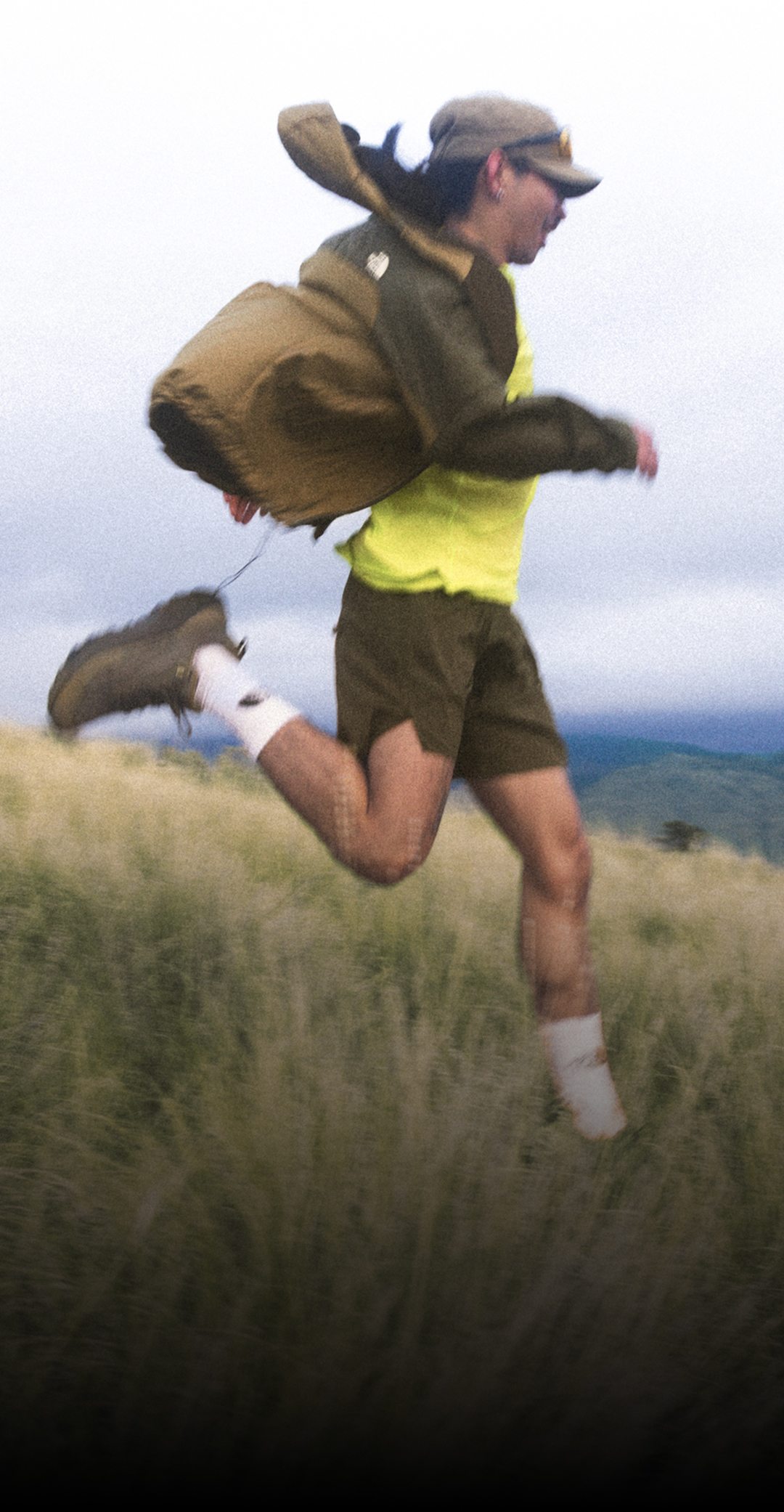 A person wearing khaki shorts, a yellow shirt, brown jacket and brown hat running joyfully down a grassy knoll.