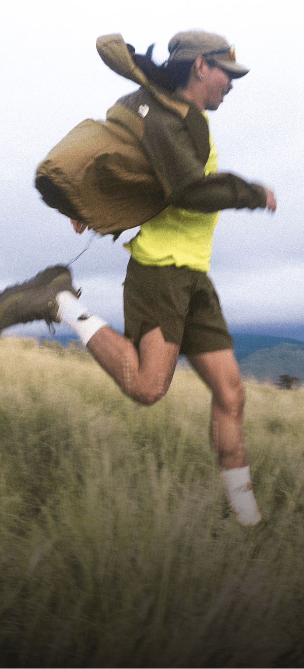 A man in a hat, khaki shorts, yellow shirt and brown jacket is running joyfully down a grassy knoll. 
