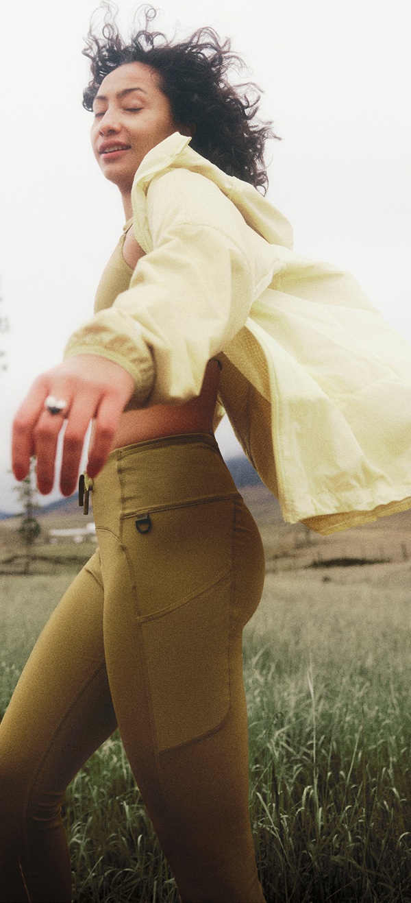 A woman in a yellow jacket and dark curly hair is twisting around. Hills and cloudy skies in the background.