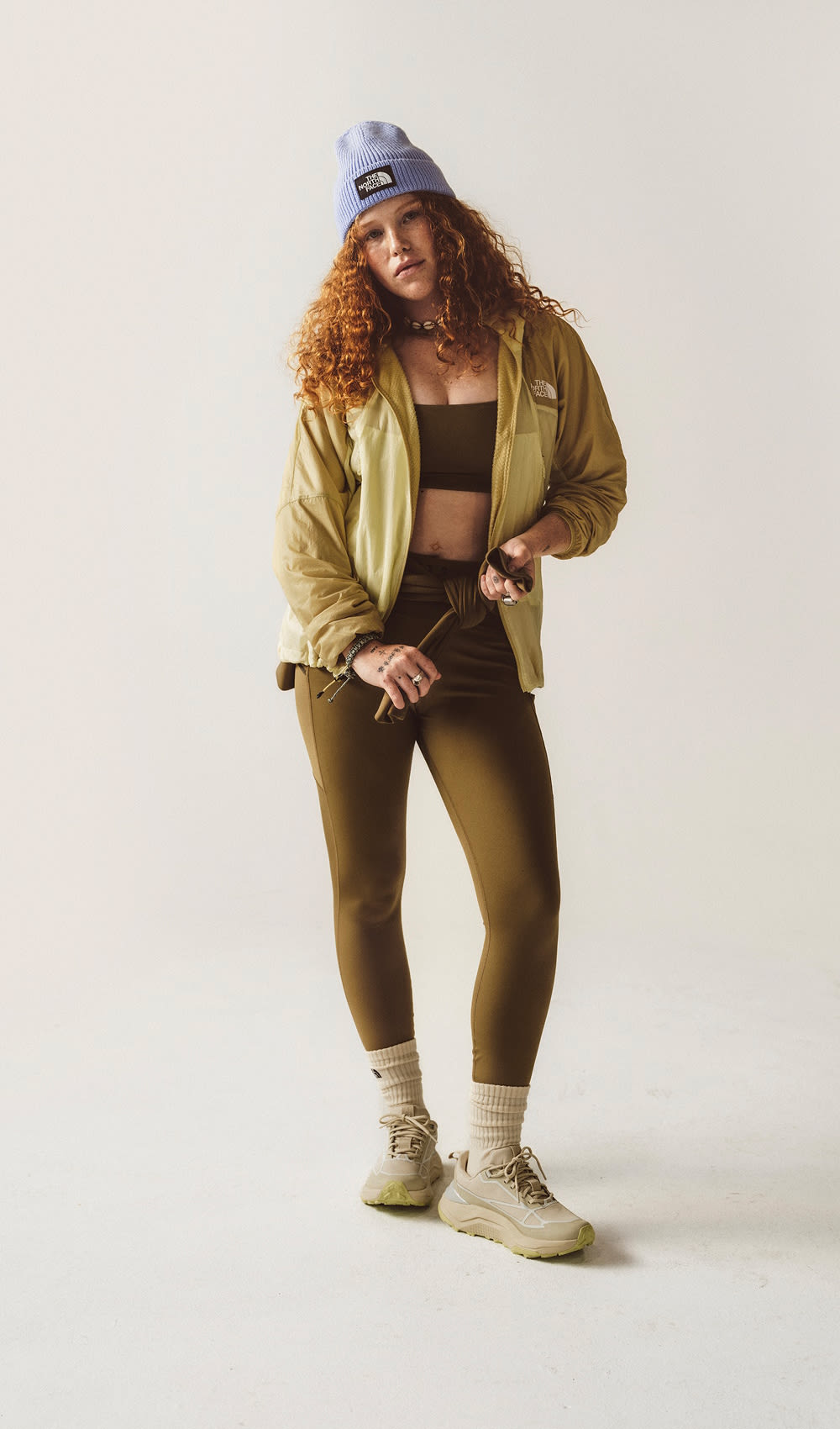 Une femme aux cheveux bouclés roux debout, portant un chapeau violet, une veste jaune et des leggings bruns.