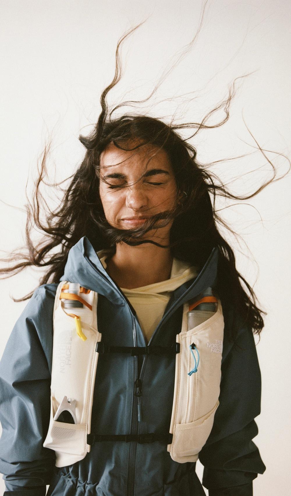 Les cheveux d’une femme balayés par le vent devant son visage, portant une veste bleue et un gilet de course blanc, sur fond blanc.