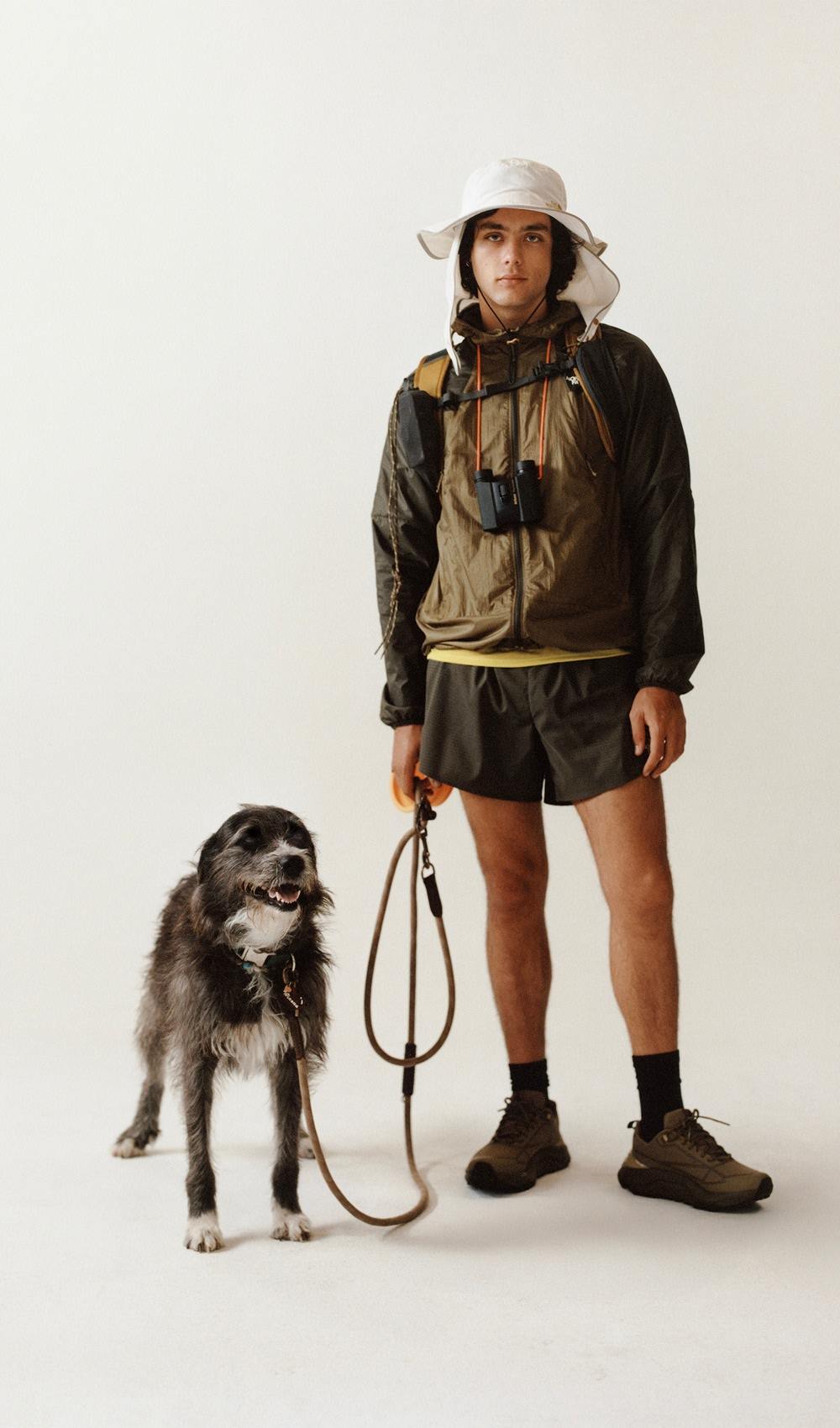 Un homme debout avec un chien, portant un chapeau, des shorts kaki, un chandail jaune et une veste brune.