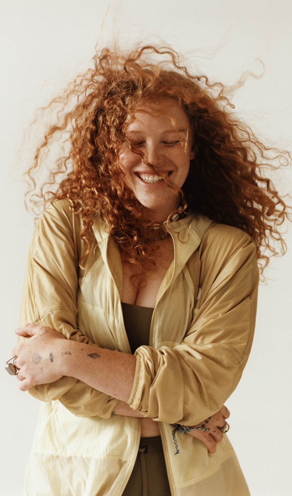 Une femme aux cheveux bouclés roux se serre dans ses bras, portant une veste jaune, sur fond blanc.