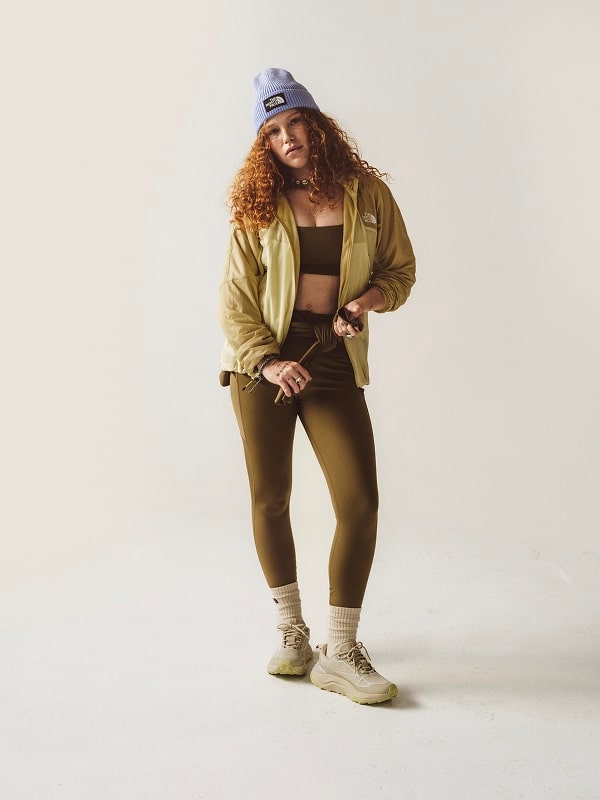 Woman with red curly hair standing wearing a purple hat, yellow jacket, and brown leggings