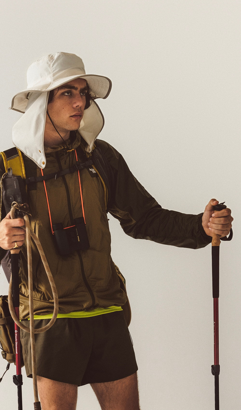 Un homme debout avec des bâtons de randonnée, regardant au loin, portant un chapeau beige, des shorts kaki, un chandail jaune et une veste brune.