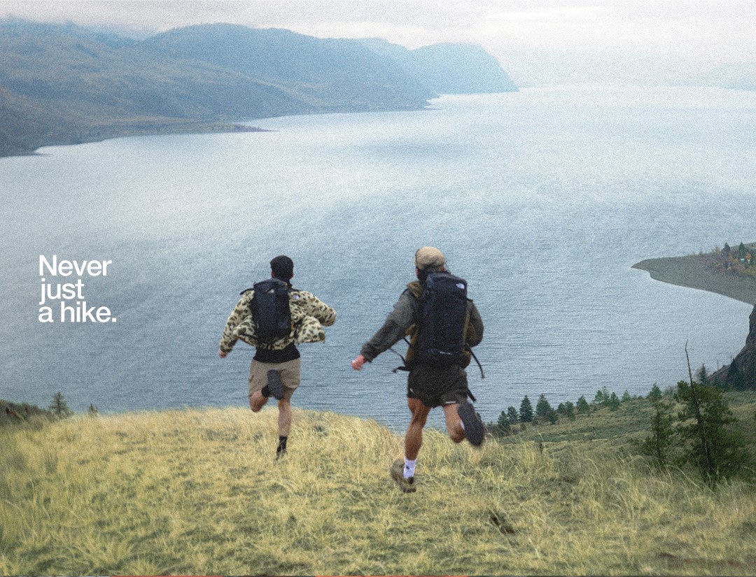Two people wearing hiking gear running down a hill in a gorge setting with overcast sky.