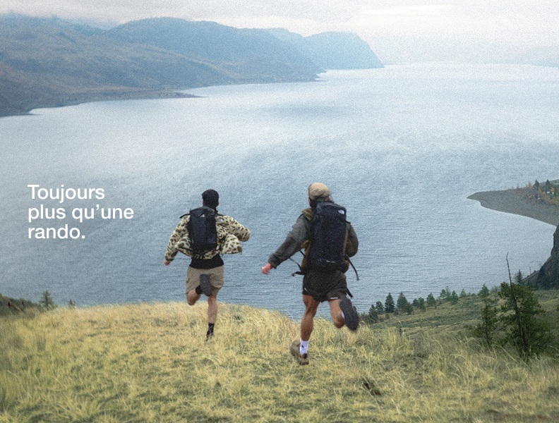 Deux personnes portant de l’équipement de randonnée courent en descendant une colline dans une gorge, sous un ciel couvert.