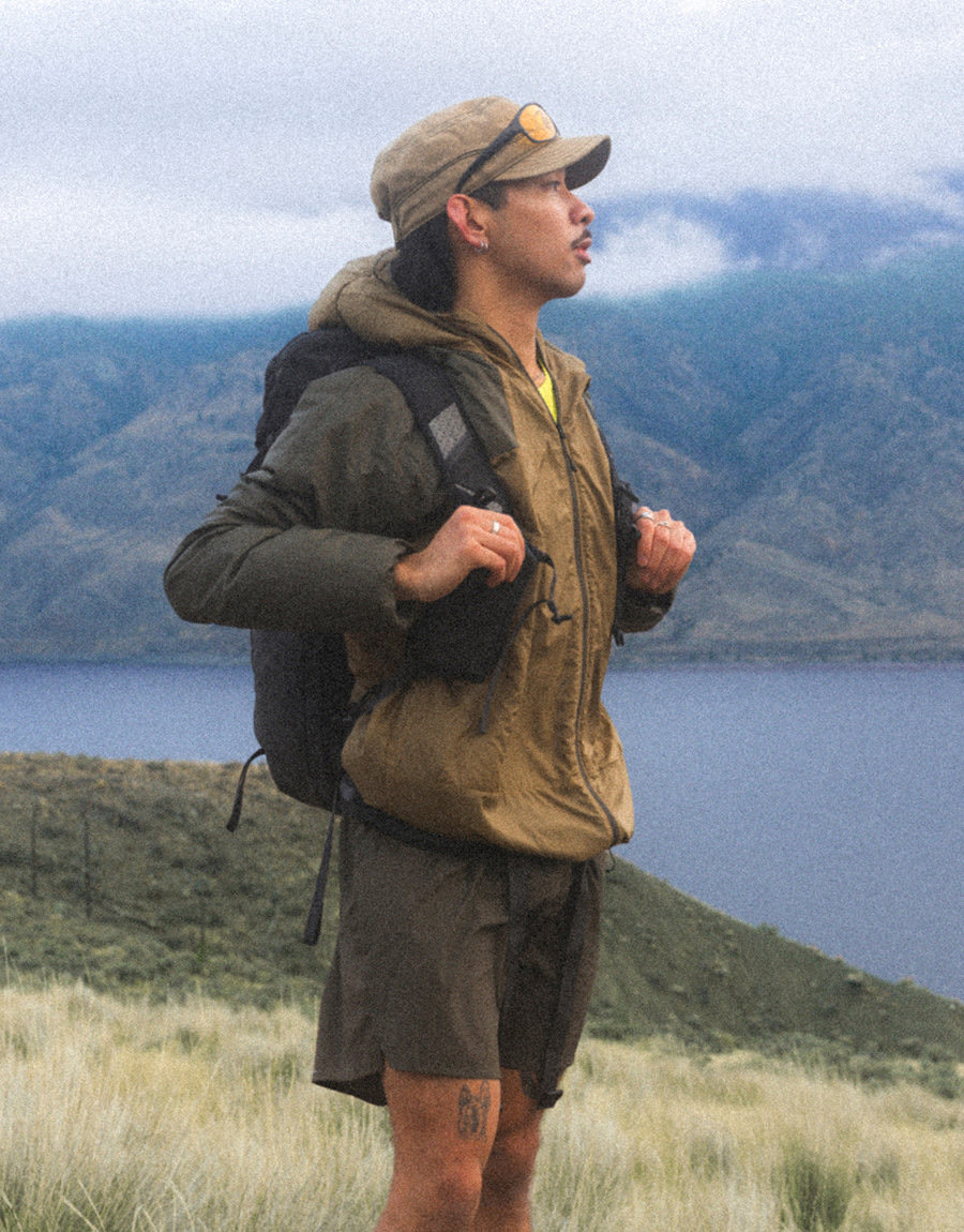 A person wearing khaki shorts, a yellow shirt, brown jacket and brown hat looking out in the distance