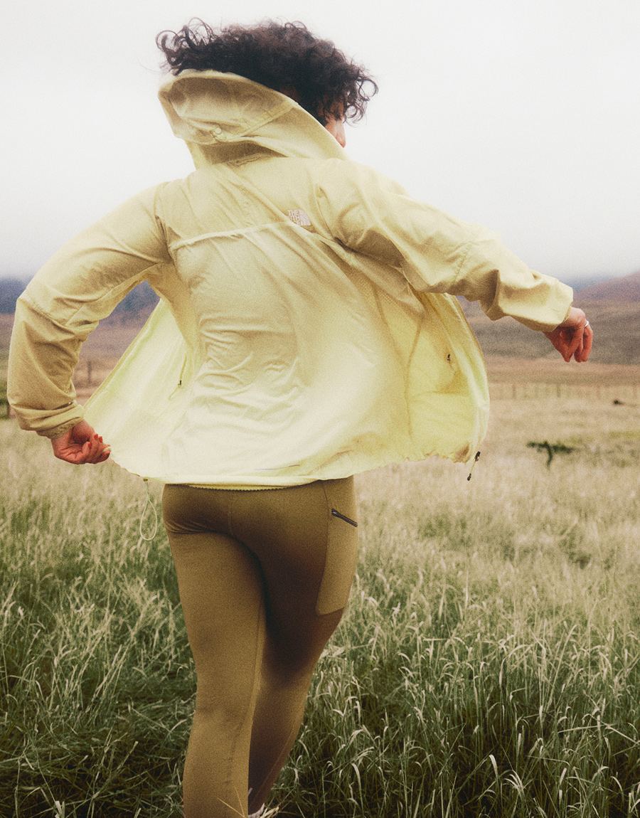 A person in a yellow jacket and dark curly hair is twisting around. Hills and cloudy skies in the background.