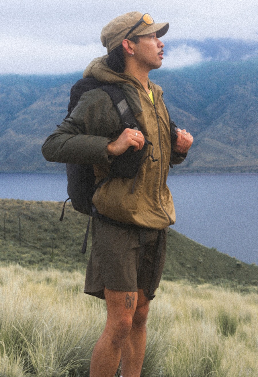 Une personne portant un short kaki, une chemise jaune, une veste brune et un chapeau brun regarde au loin.