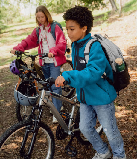Two kids walking their bikes wearing backpacks
