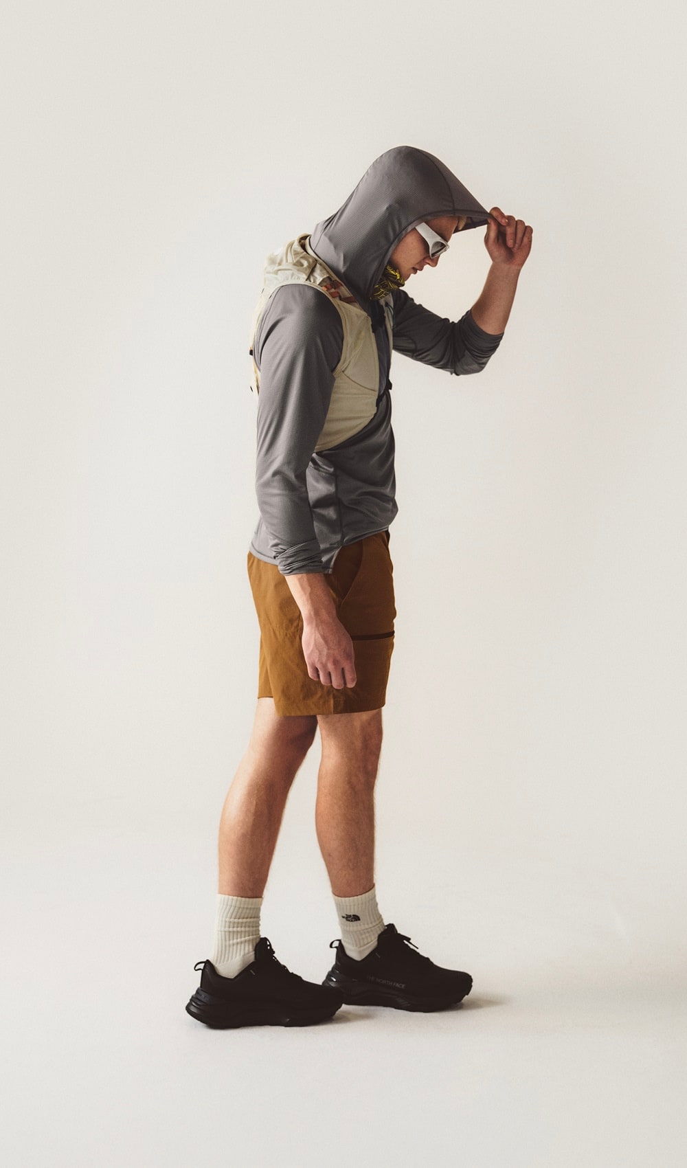 A man standing wearing a grey long sleeve with a hood with brown shorts