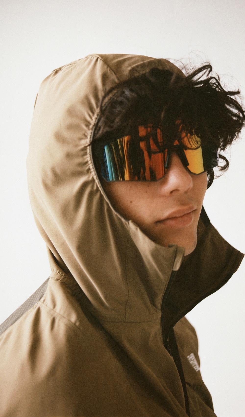 A man wearing a brown jacket with his hood up wearing sunglasses
