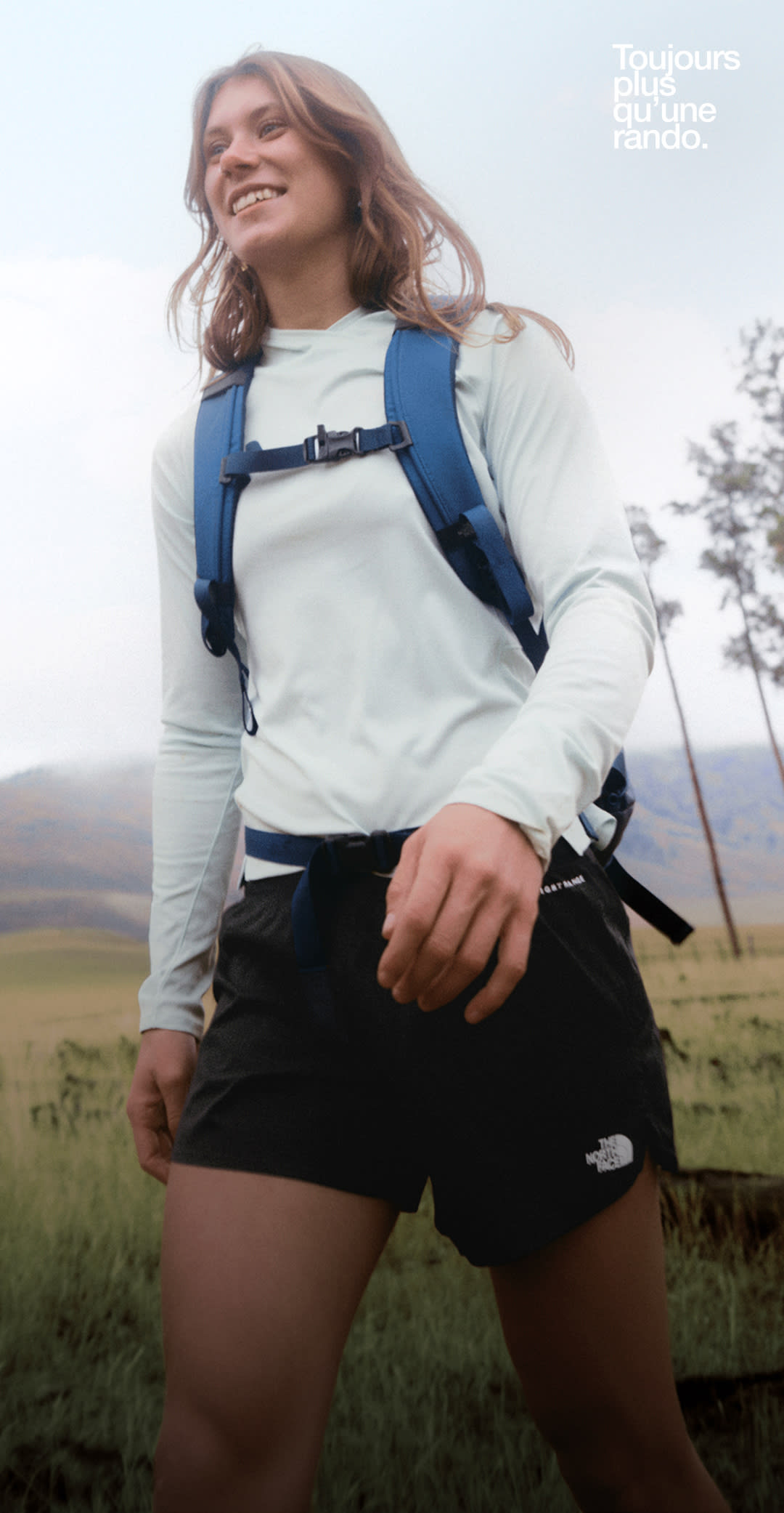 Une personne vêtue d’un chemisier bleu et portant un sac à dos bleu marche sur un terrain plat avec des collines en arrière-plan.