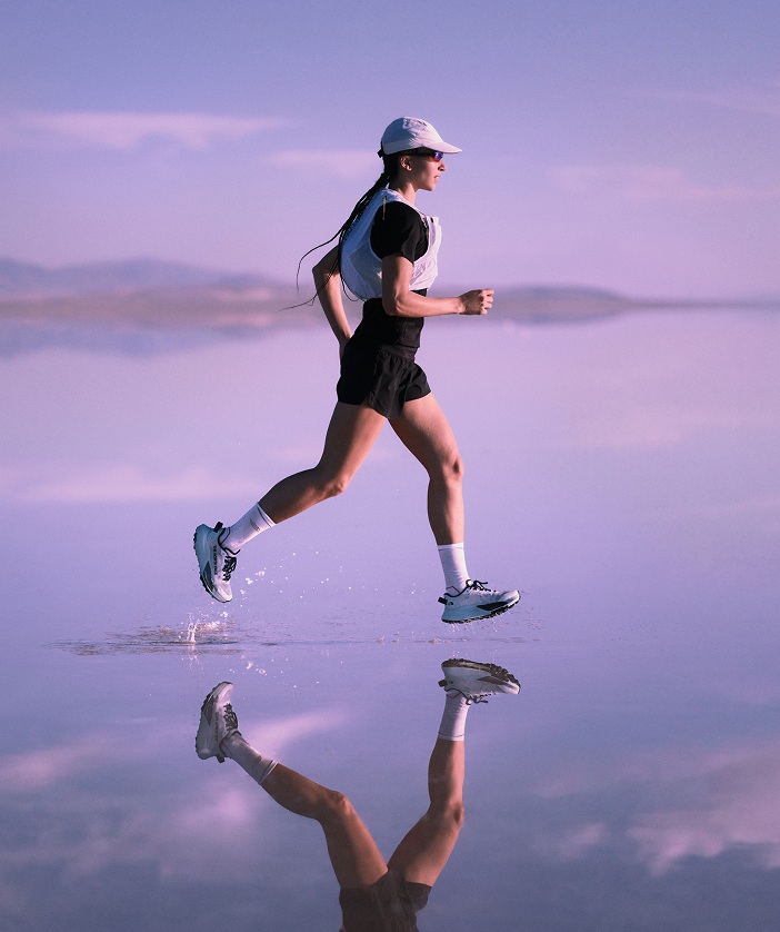 A person wearing Altamesa 500 V2 shoes running on surf, the mountains in the background reflected in the thin layer of water.