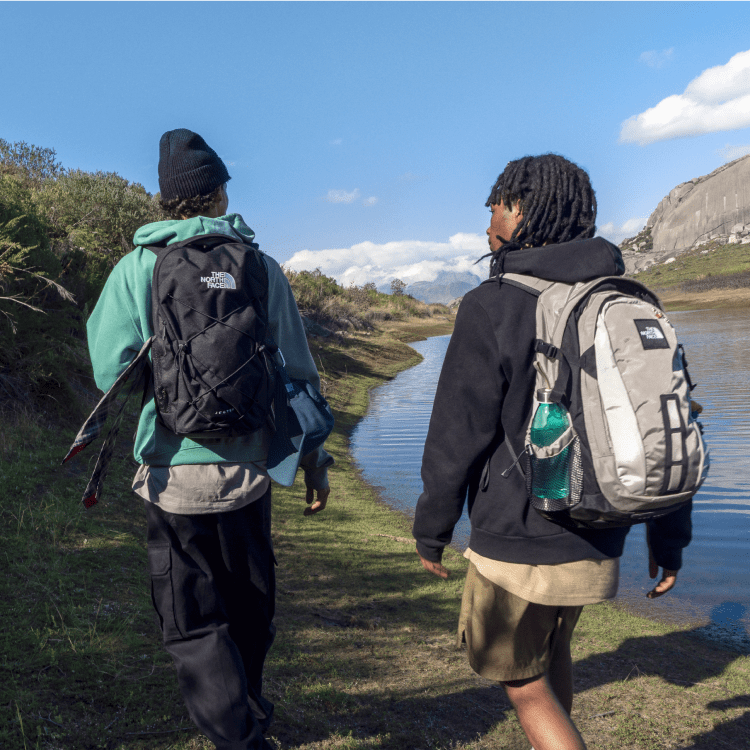 Deux jeunes portant un sac à dos, vus de dos, marchent le long d’une petite rivière.  
