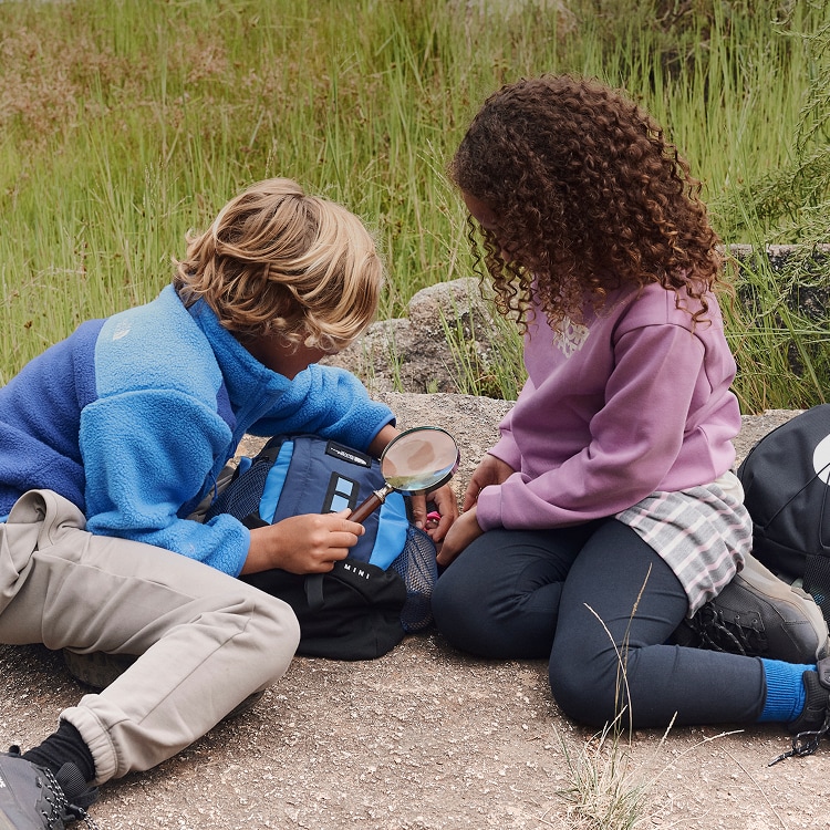 A boy wearing a blue fleece and girl wearing a purple sweatshirt looking at something through a magnifying glass.