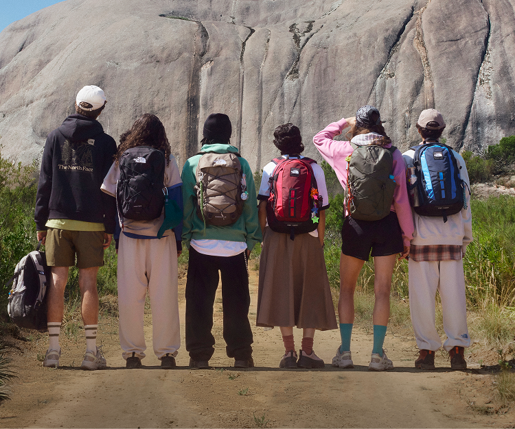 Six young people facing away from the camera, looking at a mountain in the distance. They’re all wearing backpacks. 
