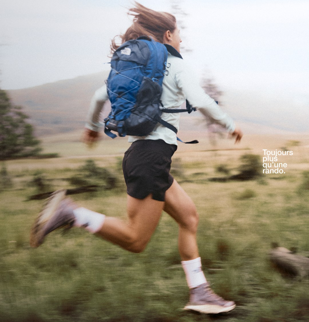 Une personne portant un short noir, une veste bleue et un sac à dos bleu, courant sur un terrain plat avec des arbres et des collines en arrière-plan.