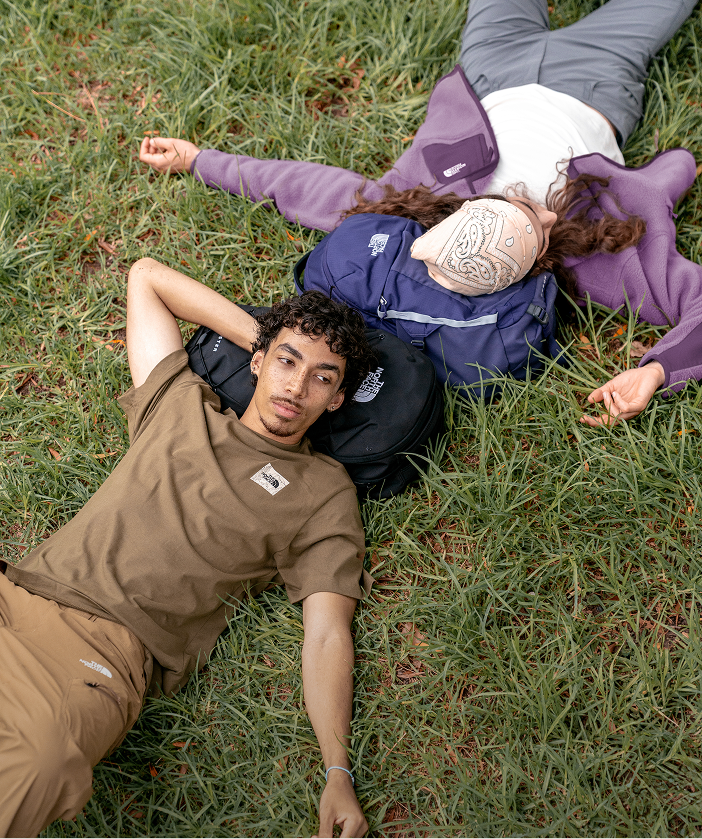 Two young people lying on the grass, heads resting on backpacks, pondering life.