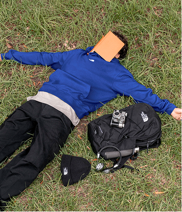 A man laying on the ground with a book covering his face, wearing a blue hoodie.
