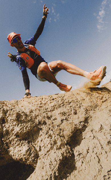 Person in orange shoes, blue shorts and orange hat running on dry mountainous terrain. 