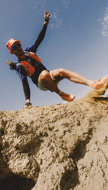 Person in orange hat and orange vest running down dry, rocky terrain.