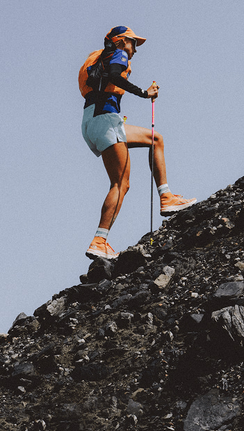 Person in orange shoes, blue shorts, orange vest and orange hat hiking up rocky terrain.