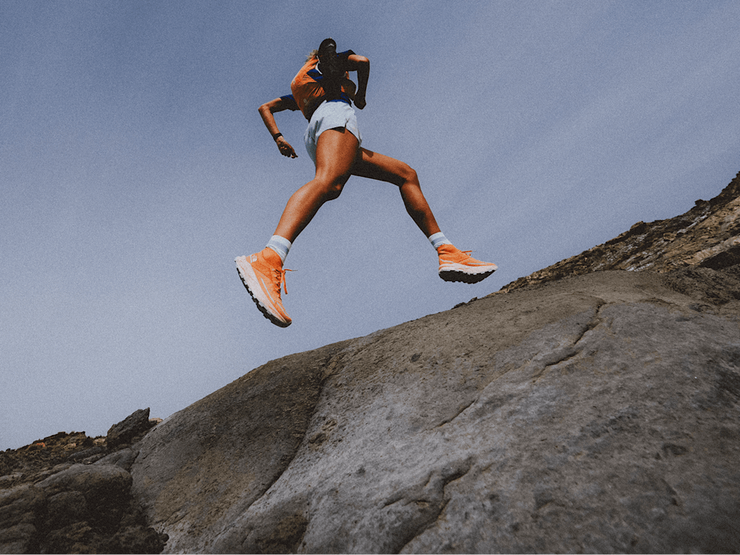 Person wearing orange shoes and blue shorts running along rocky terrain. Only the bottom half of the person is in frame, camera angle is low. 