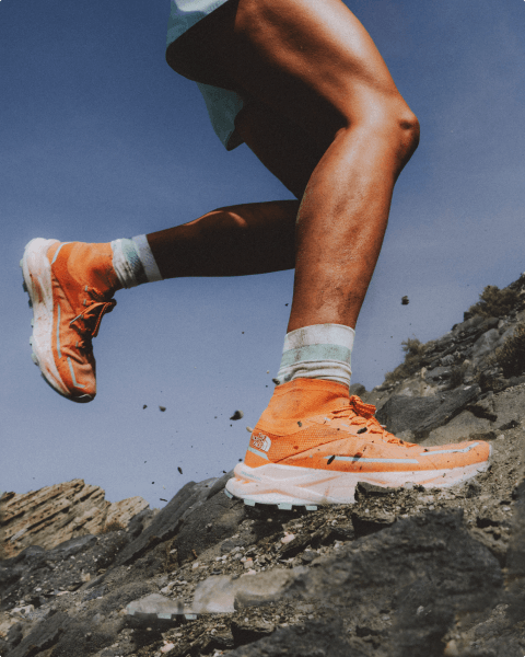 Person wearing orange shoes and blue shorts running along rocky terrain. Only the bottom half of the person is in frame, camera angle is low. 