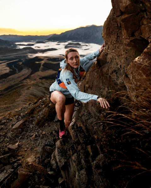 Kaytlyn Gerbin is wearing a blue jacket, orange vest and orange Offtrail Ultra shoes, climbing up a rock wall. 