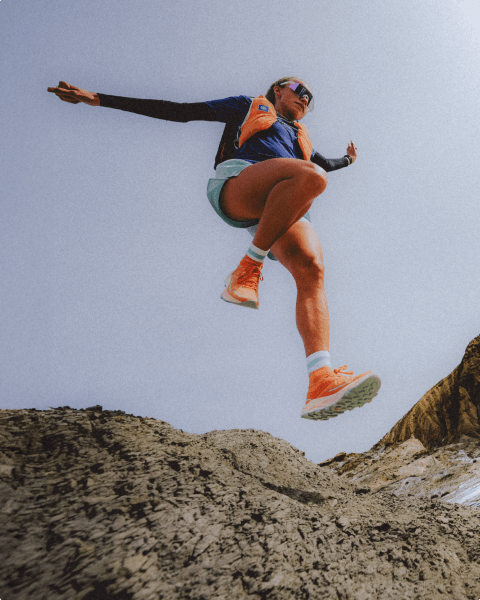 Person is bounding across a rocky surface, wearing orange Offtrail Ultra shoes, blue shorts and a blue long-sleeve shirt. 