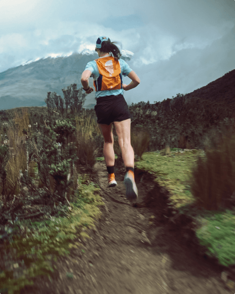 Tara Fraga is wearing a blue shirt, black shorts and orange shoes running along a dirt trail with vegetation on either side. 
