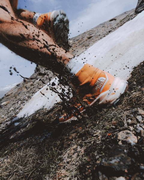 Only the person’s legs and feet are in the frame, wearing orange Offtrail Ultra shoes. They are stepping in mud, which is flying upward.
