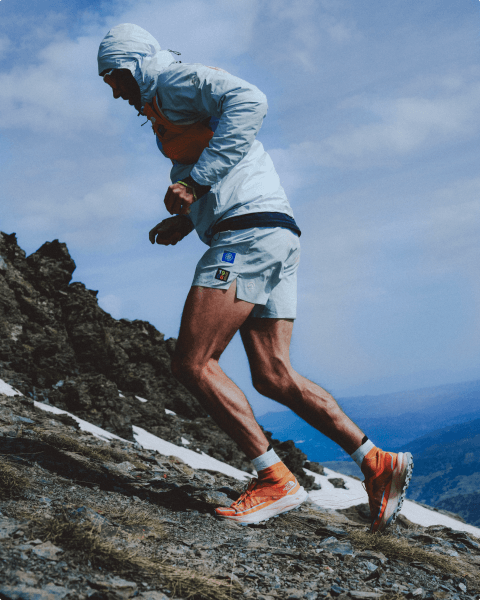 Person in blue hoodie jacket, blue shorts and orange Offtrail Ultra shoes is running up a rocky slope.