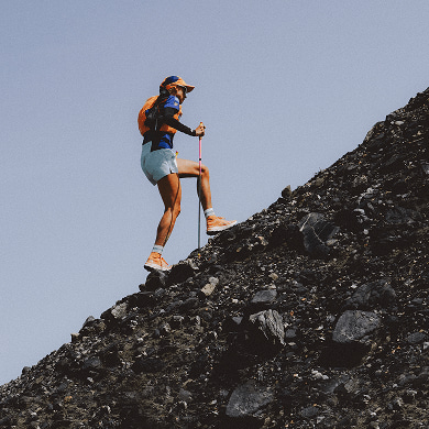 A person is hiking up a rocky slope wearing an orange Summit Series™ Run Vest.