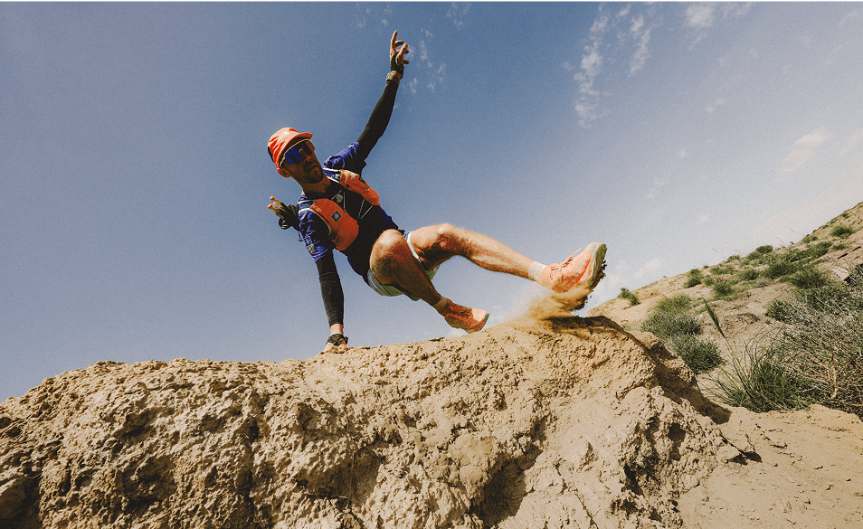 Athlete running up a dirt trail in an all-orange outfit with a black vest and running hat on.