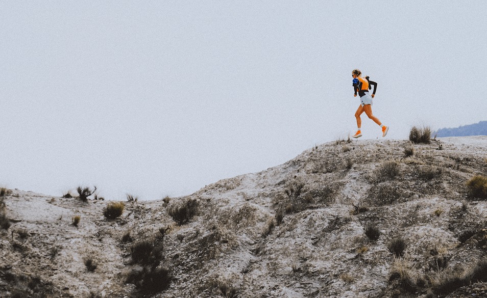 A woman running up a mountain in an orange jacket at sunset.