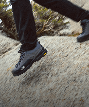 This image shows the bottom of a person’s legs wearing brown hiking pants and brown shoes, standing on arid ground. 