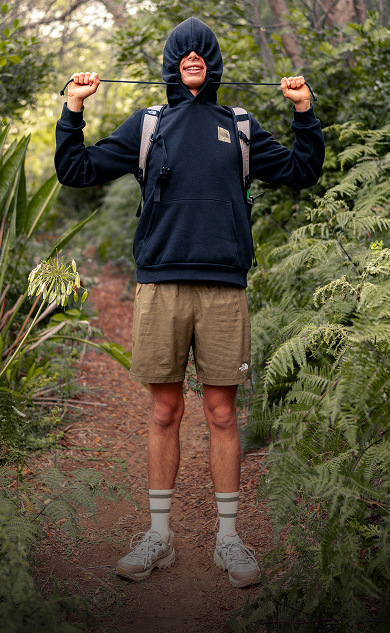 A person is standing in the forest, wearing a black hoodie with the hood pulled up and the drawstrings pulled so his face is covered.