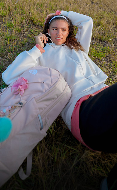 A person is standing, looking at the camera, wearing a striped shirt and light pink jacket. Trees in the background.