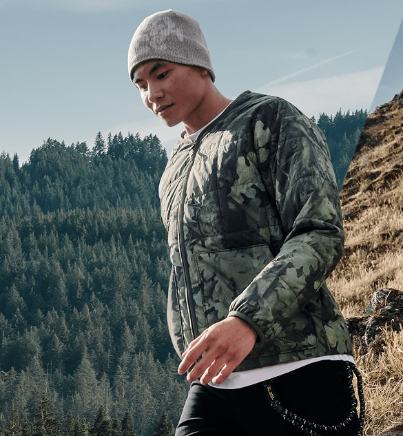 A person wearing a camouflage-patterned jacket is moving through a split screen landscape with an evergreen forest on one side and a dry grassy hillside on the other side.