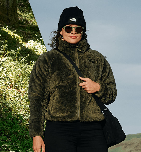 A person wearing a soft green fleece jacket is standing in a split screen landscape with an evergreen forest on one side and green grassy hills and blue sky on the other side.