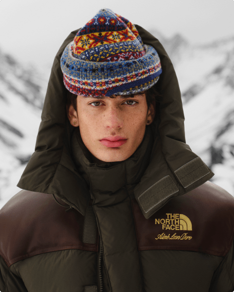 Close-up of man with jaquard knit hat and unzipped brown jacket with hood pulled up around him, with snowy mountain backdrop.