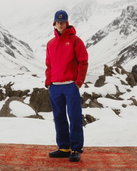 Full body shot of a man wearing a red jacket and navy pants, standing in front of a snowy mountain backdrop