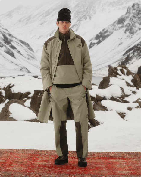 Full-body shot of man wearing an open beige trenchcoat with matching mock neck and beige pants, against a snowy mountain backdrop.   [no copy] 