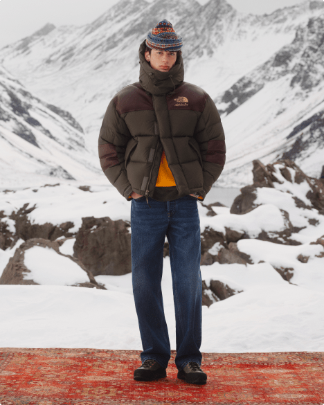 Full-body shot of a man wearing a brown zip-up puff jacket and jeans, standing against a snowy mountain backdrop.