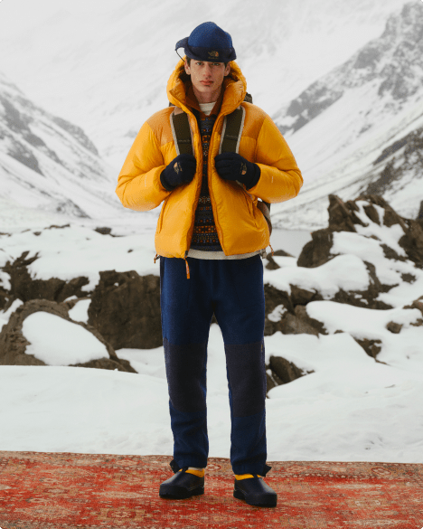 Full-body shot of man wearing a yellow jacket and navy pants, holding a backpack, standing in front of a snowy mountain backdrop.