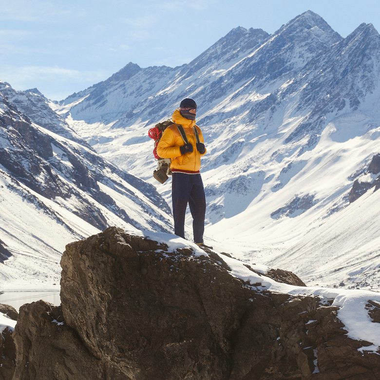 Man in yellow jacket and navy pants holding a backpack against a snowy mountain backdrop.